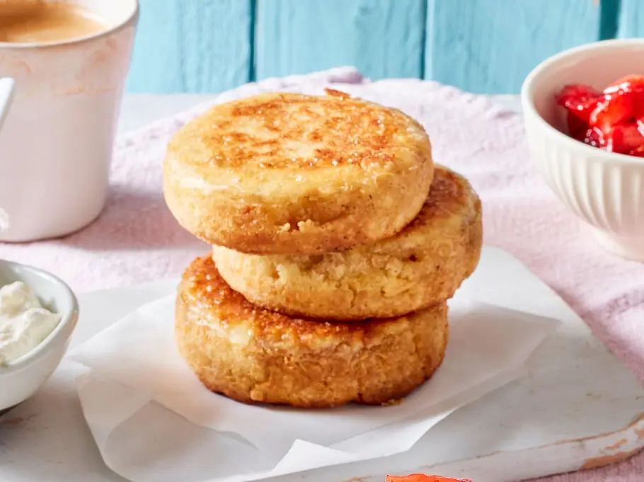 Drei goldbraun gebackene Crumpets liegen gestapelt auf einem Brett, daneben eine Ricotta-Creme und Erdbeeren in einem Schälchen