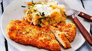 Knuspriges Schnitzel in goldbrauner Brezelpanade, serviert mit bayerischem Kraut und Kräuterschmand auf einem weißen Teller, dazu Besteck auf hellem Holztisch. - Foto: LECKER @ Bauer Media Group Knuspriges Schnitzel in goldbrauner Brezelpanade, serviert mit bayerischem Kraut und Kräuterschmand auf einem weißen Teller, dazu Besteck auf hellem Holztisch. - Foto: LECKER @ Bauer Media Group