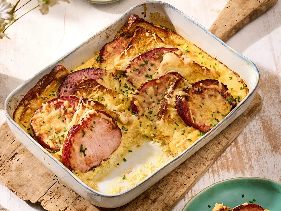 Kasseler-Brot-Auflauf Ein Auflauf mit Brot, Fleischscheiben, Käse und Schnittlauch steht auf einem Holzbrett. Daneben stehen ein portionierter Teller und Bier.