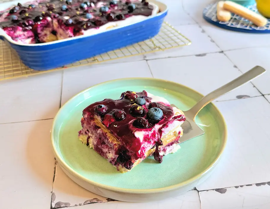 Heidelbeer-Tiramisu Rezept Ein Stück Heidelbeer-Tiramisu mit Beeren und Creme liegt auf einem Teller, daneben eine Auflaufform mit restlichem Dessert.