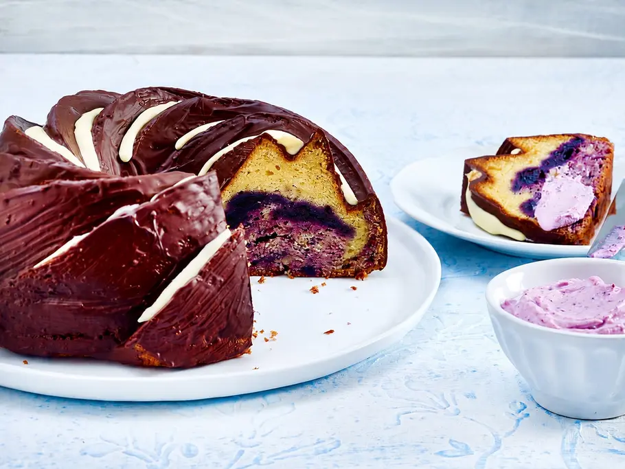 Heidelbeer-Donauwelle Rezept Eine angeschnittene Heidelbeer-Donauwelle mit Schokoglasur steht neben einem Teller mit Kuchenstück und einer Schale lila Creme.