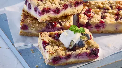 Heidelbeer-Streuselschnitten mit knusprigen Streuseln und frischen Heidelbeeren auf Backpapier, eine Schnitte mit Sahne und Beeren garniert, angerichtet auf einem hellblauen Holzbrett. - Foto: LECKER @ Bauer Media Group