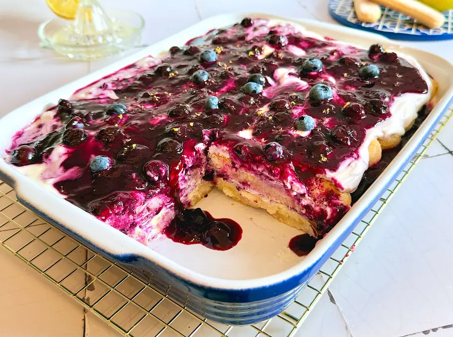 Eine Auflaufform mit angeschnittenem Heidelbeer-Tiramisu, getoppt mit frischen Heidelbeeren und Zitronenabrieb.