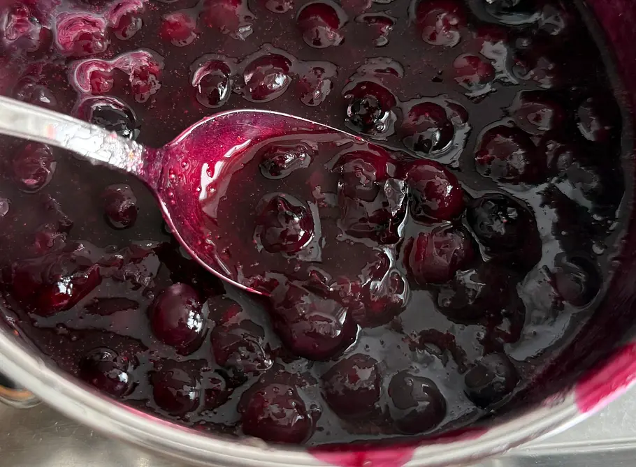  In einem Topf liegt ein Löffel in dunklem Heidelbeerkompott mit ganzen, glänzenden Heidelbeeren.