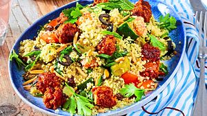 Bunter Couscous-Salat mit Gemüse, frischen Kräutern, Oliven und knusprigen Fleischbällchen auf einem blauen Teller angerichtet. - Foto: Image Professionals