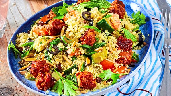 Bunter Couscous-Salat mit Gemüse, frischen Kräutern, Oliven und knusprigen Fleischbällchen auf einem blauen Teller angerichtet. - Foto: Image Professionals