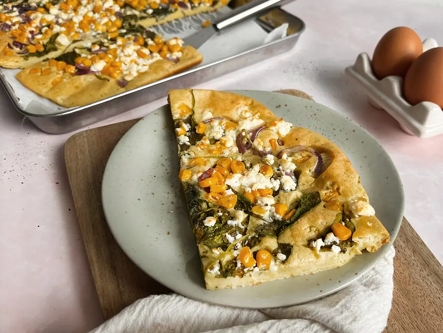 Herzhafter Pfannkuchen vom Blech Ein Stück herzhafter Pfannkuchen mit Spinat, Feta und Mais auf einem grünen Teller und Holzbrett, im Hintergrund Eier und Blech mit Rest Pfannkuchen