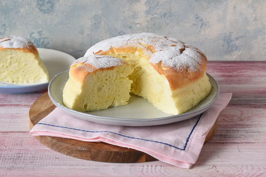 High-Protein Wölkchenkuchen: Das virale Rezept ohne Zucker Ein angeschnittener, mit Puderzucker bestäubter Kuchen liegt auf einem Teller mit rosa Serviette auf einem Holzbrett.