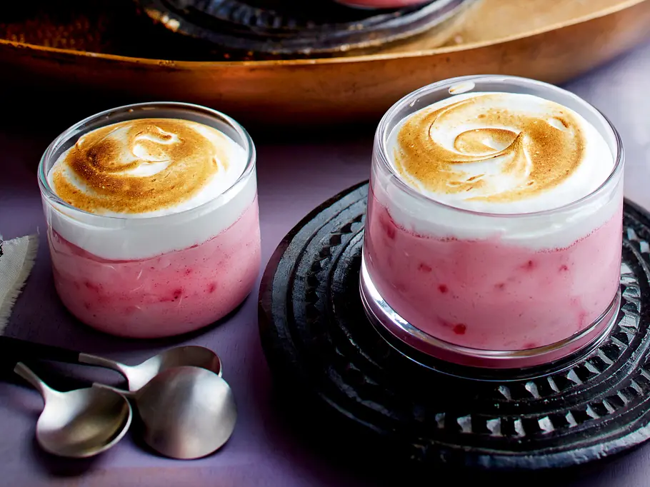 Himbeer-Baiser-Creme Rezept Zwei Gläser mit rosafarbener Himbeercreme, darauf goldbraun gebräuntes Baiser, eins steht auf einem dunklen Untersetzer.