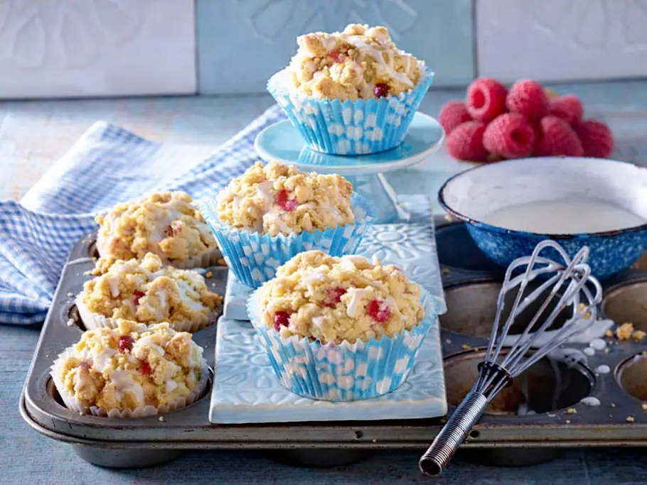 Sechs Himbeer-Muffins mit Streuseln auf einem Muffinblech. Im Hintergrund liegen ein paar frische Himbeeren als Deko.