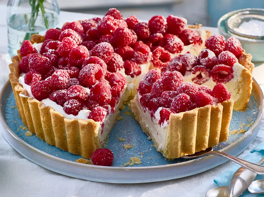 Himbeer-Ricotta-Tarte auf einem hellblauen Teller, mit frischen Himbeeren und Puderzucker bestreut, auf weißer Tischdecke serviert.