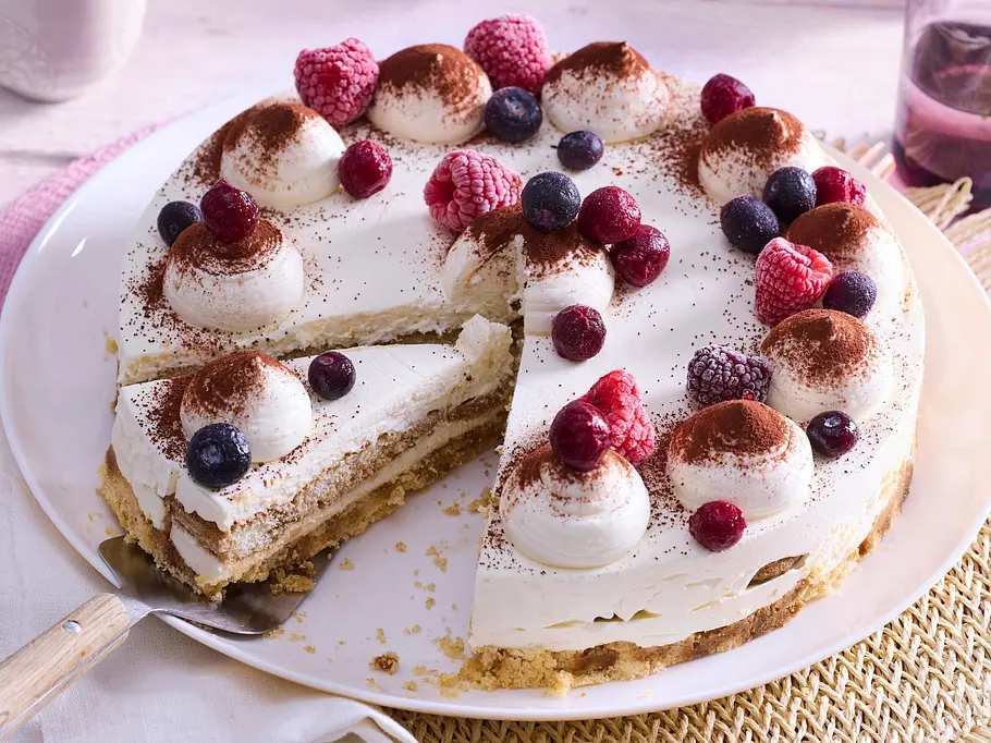 Eine angeschnittene Tiramisu-Torte ist mit Sahnetupfen, Kakao und verschiedenen Beeren dekoriert.
