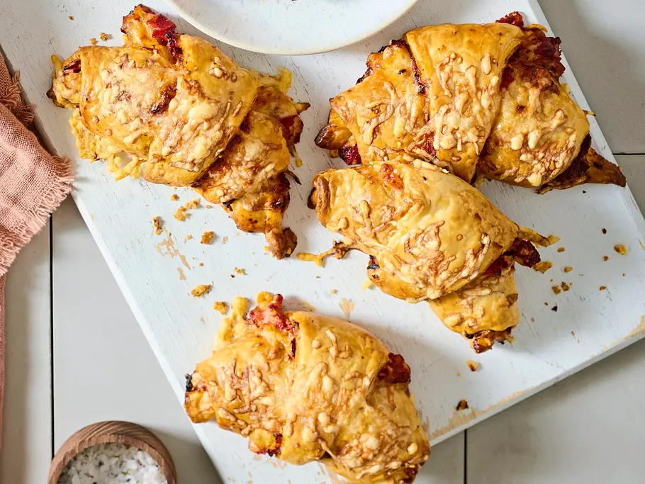 Goldbraune Croissants mit geschmolzenem Käse und Pizzabelag, serviert auf einem weißen Holzbrett und einem kleinen Teller, daneben ein brauner Salznapf und ein beigefarbenes Tuch auf heller Kachelunterlage.