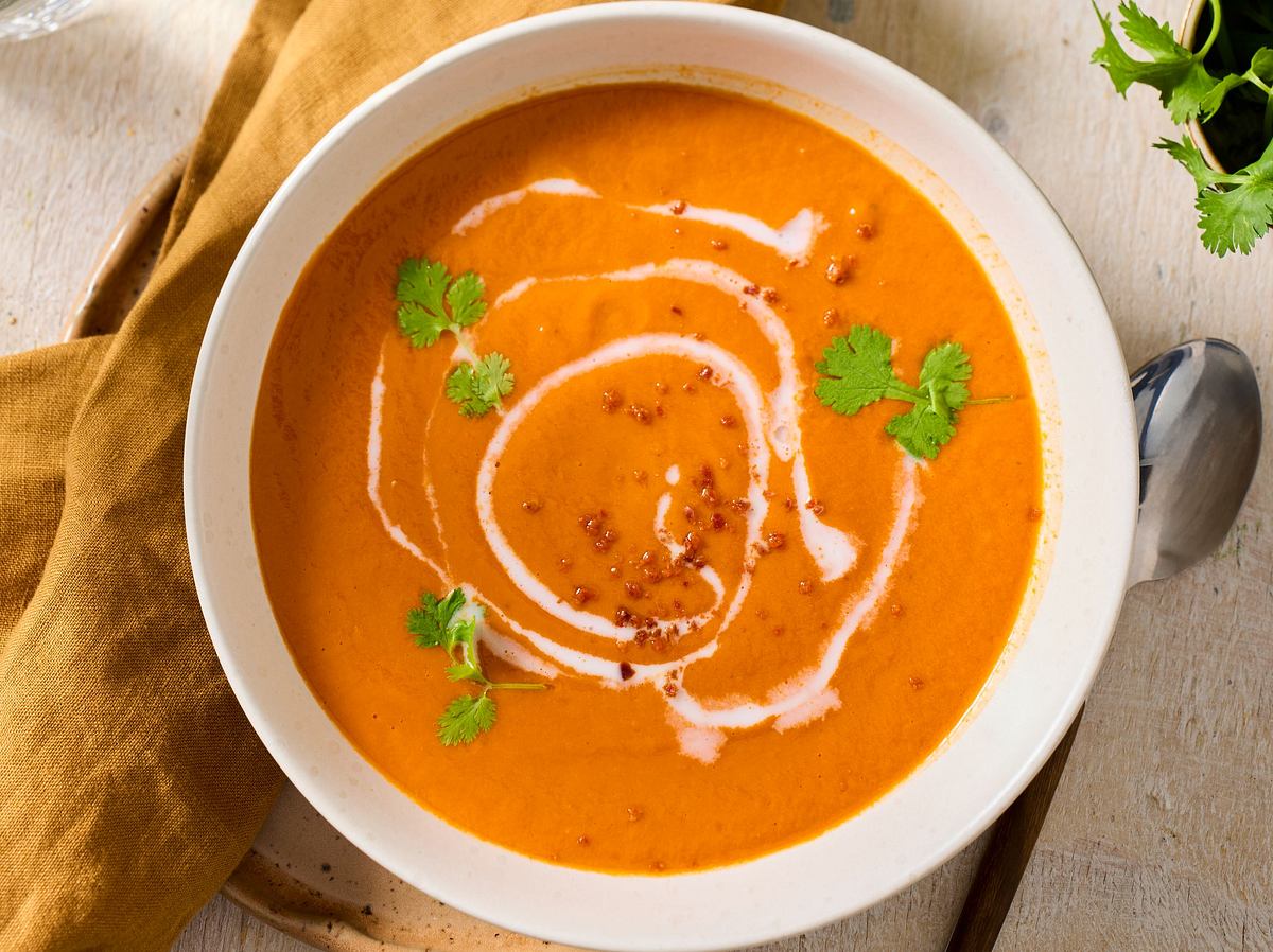 Cremige Tomatensuppe mit Kokosmilch, garniert mit frischem Koriander und einem Swirl aus Kokosmilch, serviert in einer weißen Schale auf einem hellen Holztisch mit gelbem Stoffserviette.