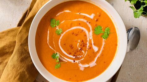 Cremige Tomatensuppe mit Kokosmilch, garniert mit frischem Koriander und einem Swirl aus Kokosmilch, serviert in einer weißen Schale auf einem hellen Holztisch mit gelbem Stoffserviette. - Foto: LECKER @ Bauer Media Group