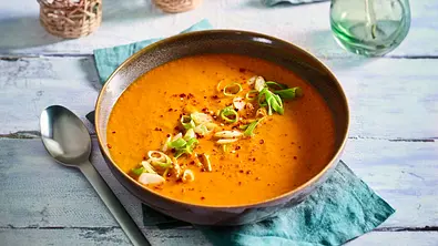 Cremige Röstpaprika-Suppe in einer braunen Schale, garniert mit Frühlingszwiebeln und Gewürzen, auf einem türkisfarbenen Stoffserviette serviert. - Foto: LECKER @ Bauer Media Group