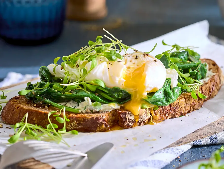 Geröstete Brotscheibe belegt mit frischem Blattspinat, pochiertem Ei mit aufgeschnittenem Eigelb und Kresse, serviert auf einem weißen Backpapier auf rustikalem Holztisch.