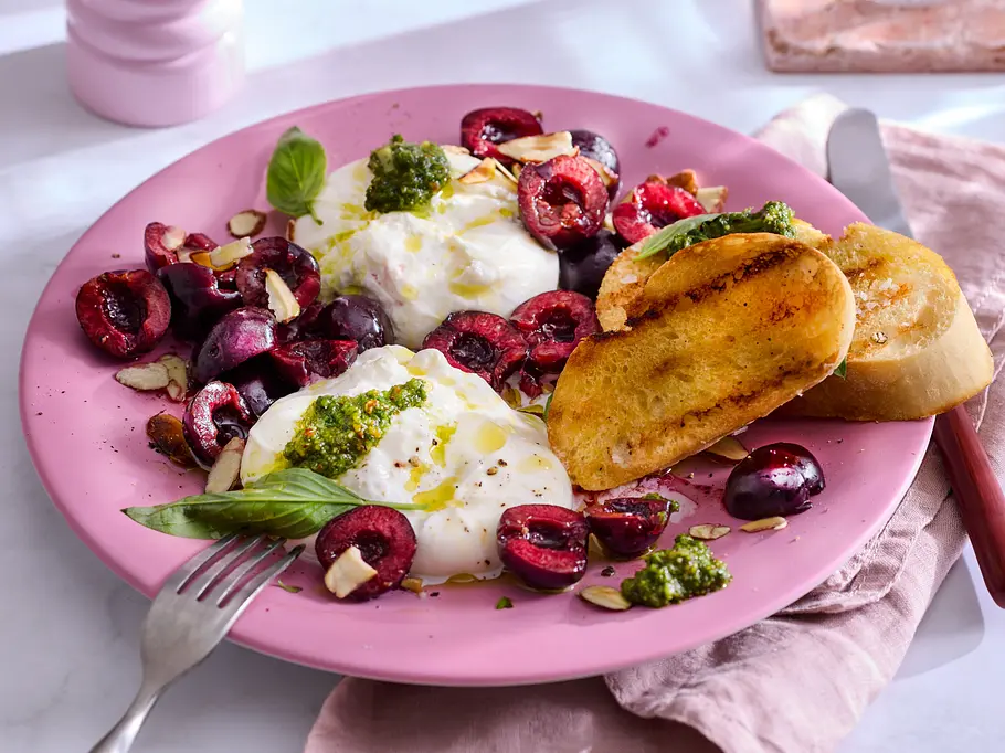 Cremige Burrata, serviert mit marinierten Kirschen, frischem Basilikum, Pesto und gerösteten Brotscheiben auf einem rosa Teller.