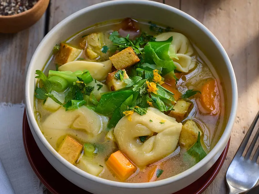 Tortellinisuppe mit herbstlichem Gemüse wie Karotten, Lauch und Tofu, serviert in einer weißen Schale auf einem Holztisch.