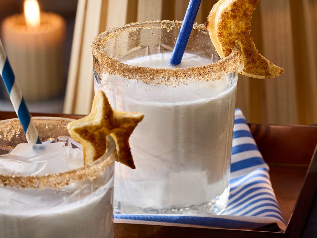 Cremiger, heller Cocktail „Geheimnisvolle Rita“ im Glas mit Zimt-Zuckerrand, Eiswürfeln, Trinkhalm und knusprigem Gebäck in Stern- oder Mondform am Glasrand.