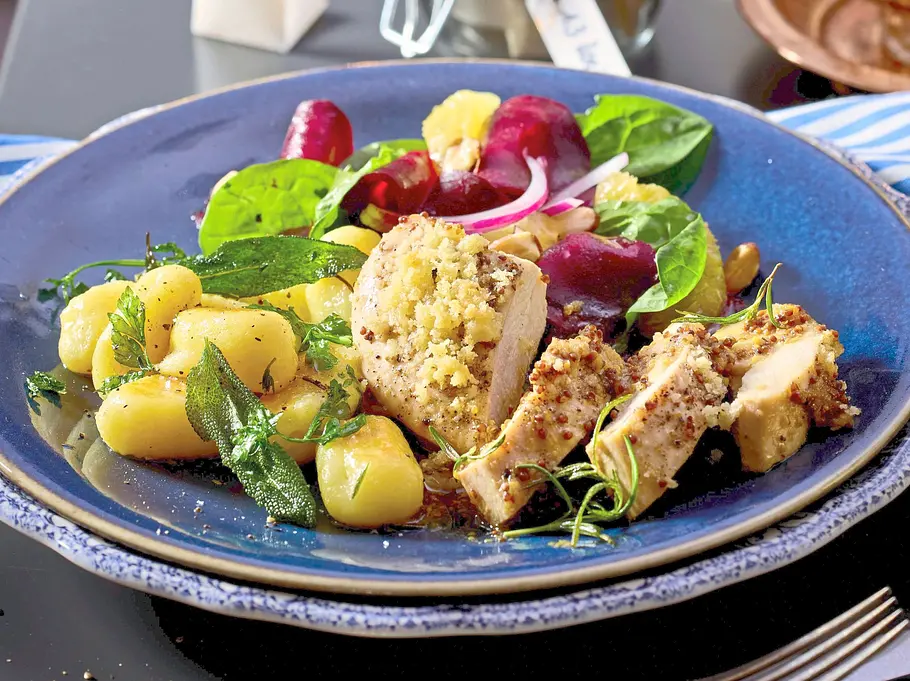 Honig-Ofenhähnchen in Scheiben, serviert mit goldenen Kräuter-Gnocchi und buntem Salat auf einem blauen Keramikteller. Daneben ein rustikales Glas und Besteck auf dunkler Tischplatte.