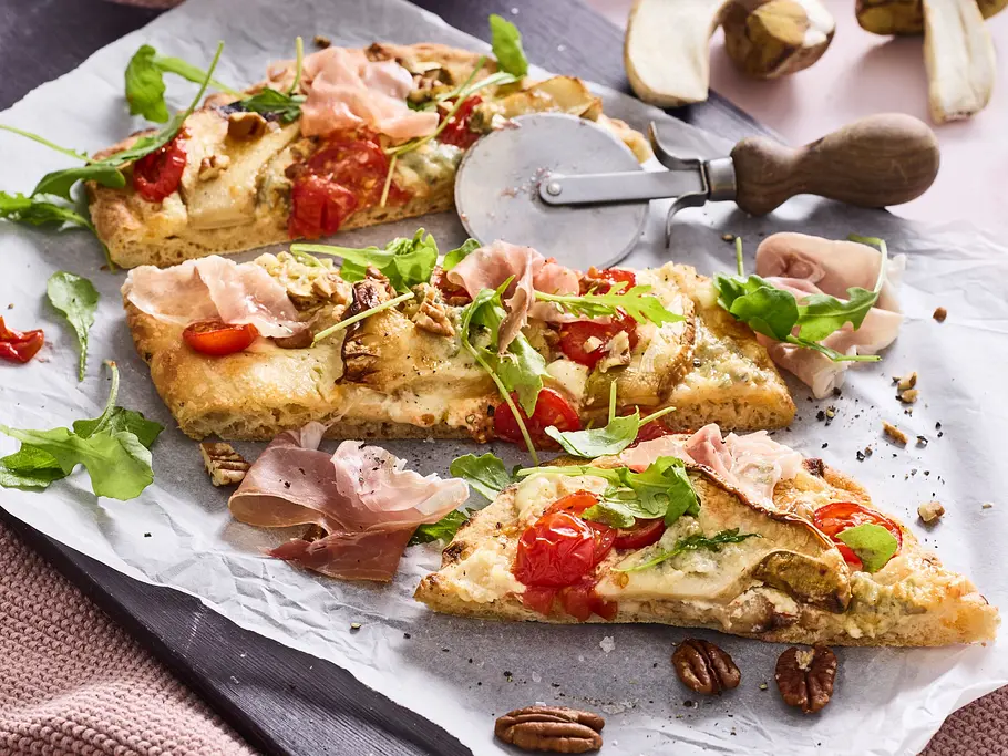 Pinsa mit Steinpilzen und Gorgonzola Aufgeschnittene Pinsastücke mit Pilzen, Tomaten, Rucola und Schinken auf Backpapier, angerichtet auf einem dunklen Holzbrett, daneben eine kleine Schale mit Pekannüssen auf rosa Unterlage.