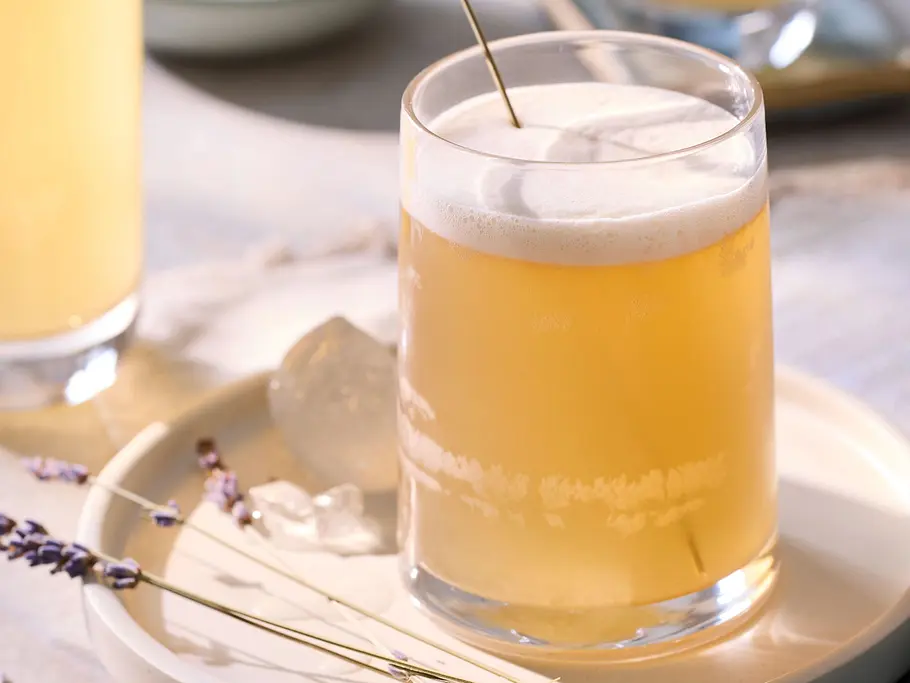 Cremiger, schaumiger Earl Grey Lavendel Drink in einem Glas mit Eiswürfeln, garniert mit einem Lavendelzweig, serviert auf einem weißen Teller mit weiteren Lavendelblüten und blauem Serviertuch.