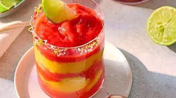 Geschichtetes Getränk aus leuchtend rotem Erdbeer- und gelbem Mango-Püree in einem Glas mit Salz-Chili-Rand, garniert mit einer frischen Limettenscheibe, serviert auf einem weißen Unterteller. - Foto: LECKER @ Bauer Media Group