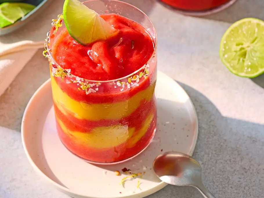 Geschichtetes Getränk aus leuchtend rotem Erdbeer- und gelbem Mango-Püree in einem Glas mit Salz-Chili-Rand, garniert mit einer frischen Limettenscheibe, serviert auf einem weißen Unterteller.