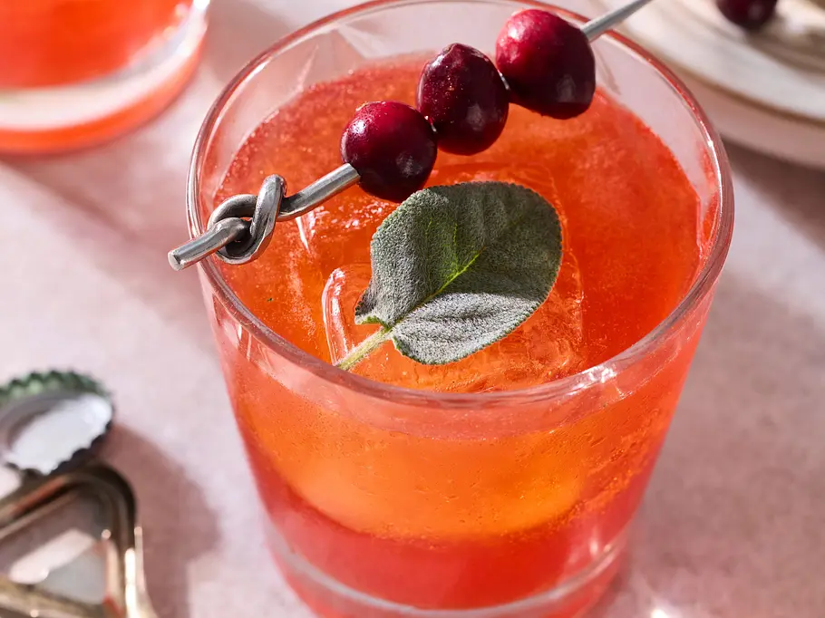 Roter Cranberry Negroni in einem kurzen Glas mit großen Eiswürfeln, garniert mit frischen Cranberries am Spieß und einem grünen Blatt, serviert auf einer hellen Unterlage.