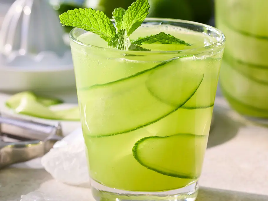 Erfrischender Gurke-Minz-Drink in einem Glas mit Eiswürfeln, dekoriert mit Minzblättern und dünnen Gurkenscheiben, serviert auf einer hellen Unterlage.