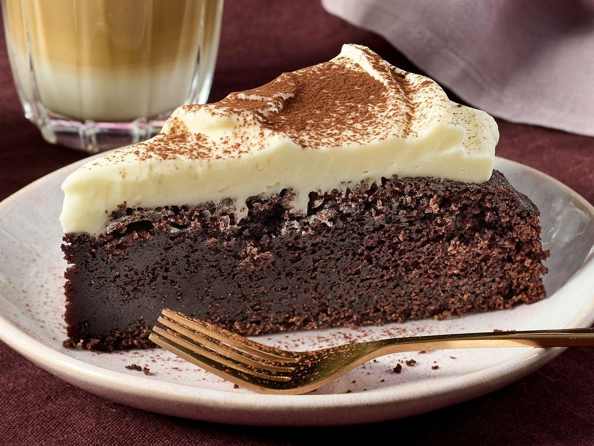 Dichter, dunkler Schokoladenkuchen mit Guinness, bedeckt mit einer dicken Schicht cremigem, weißem Frosting und leicht mit Kakaopulver bestäubt. Ein Stück des Kuchens liegt auf einem Teller davor.