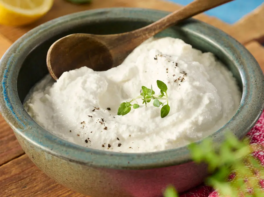 Schneller „Ricotta“ alla casa Rezept Cremiger, frischer Ricotta in einer Schale angerichtet, garniert mit schwarzem Pfeffer und frischen Kräutern. Ein Holzlöffel liegt auf der Schale.