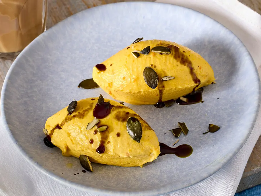 Kürbismousse Auf zwei Tellern liegen gelbe Nocken mit Kernen und dunkler Sauce. Daneben stehen zwei Gläser Kaffee und eine Blumenvase.