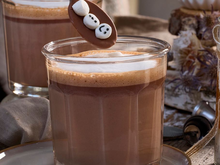 Heiße, cremige Schokolade in einem schlichten Glas, serviert auf einer Untertasse, dazu ein Schokolöffel mit Schneemann-Dekor.