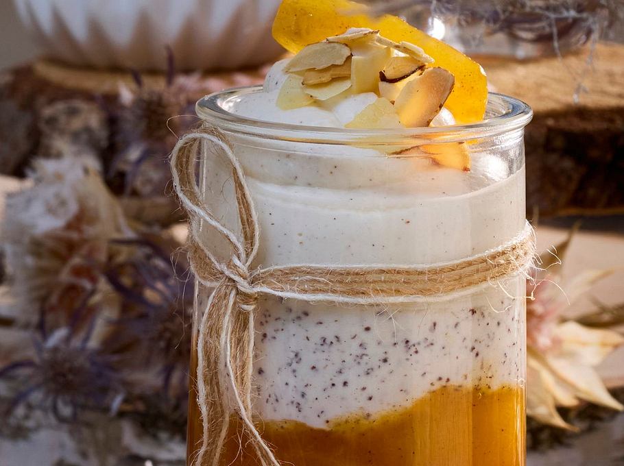 Schichtdessert aus cremigem Topfen mit Mohn und fruchtigem Aprikosenröster, angerichtet in einem hohen Glas auf rustikalem Holztisch, garniert mit gehobelten Mandeln und Orangenfilet.