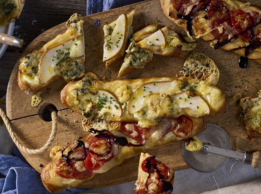 Knuspriger Rahmfladen auf Holzbrett, belegt mit Tomaten, Salami, Birnenscheiben und Kräutern, goldbraun gebacken und abwechslungsreich garniert.