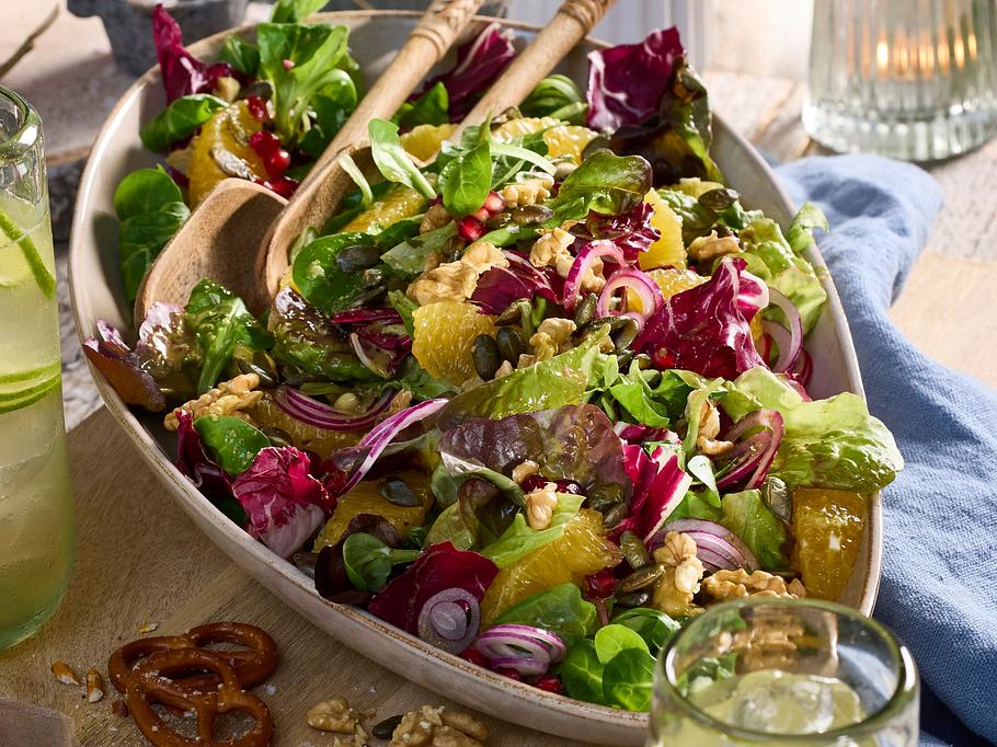 Bunter Alpensalat mit Blattsalaten, Orangenfilets, Granatapfelkernen, Walnüssen und roten Zwiebeln in einer ovalen Schale auf einem Holzbrett angerichtet.