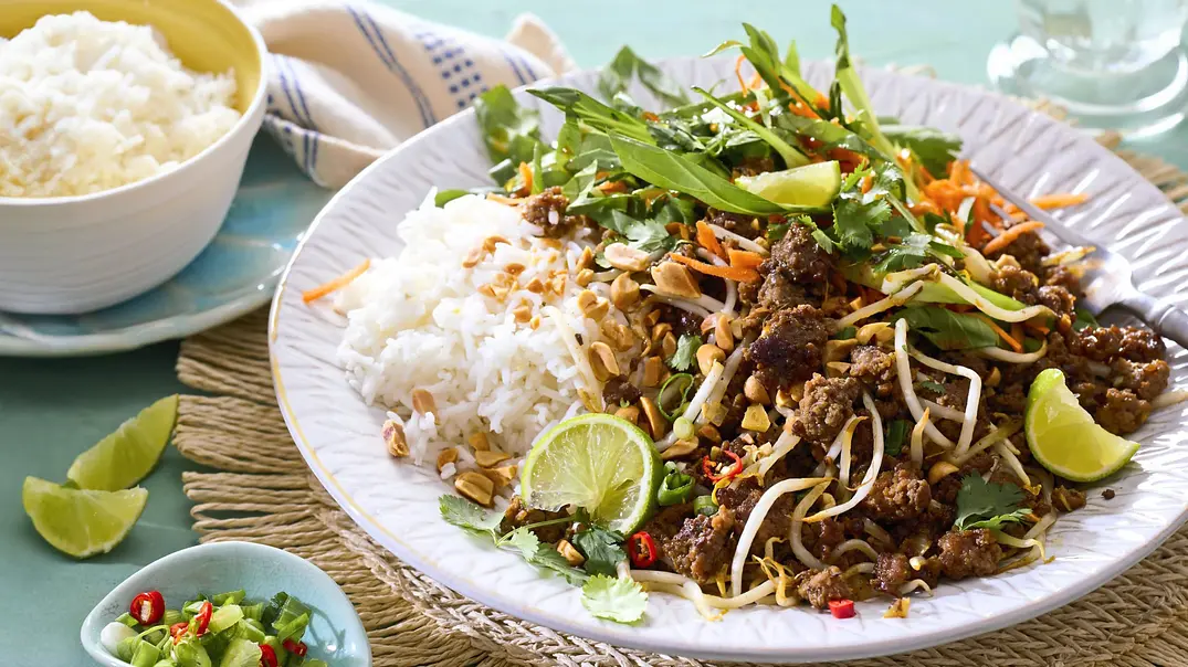 Würzig gebratenes Hackfleisch mit Limette, Sprossen, Karottenstreifen, frischen Kräutern und gerösteten Erdnüssen, serviert mit lockerem Duftreis auf einem weißen Teller mit geflochtener Unterlage. - Foto: LECKER @ Bauer Media Group