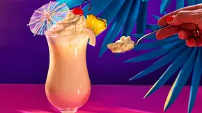 Cremige Piña-Colada-Mousse im Cocktailglas, garniert mit einem Klecks Sahne, einer Ananasscheibe, einer Cocktailkirsche und einem bunten Papier-Schirmchen. Ein Löffel mit der Mousse daneben. - Foto: LECKER @ Bauer Media Group