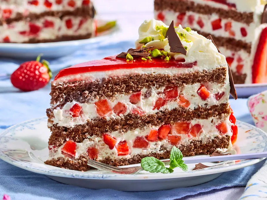 Stück festliche Erdbeertorte mit lockeren Schokobiskuit-Schichten, Sahnecreme und frischen Erdbeerstückchen, garniert mit Schokolade, Pistazien und Sahnetupfen auf einem weißen Teller.