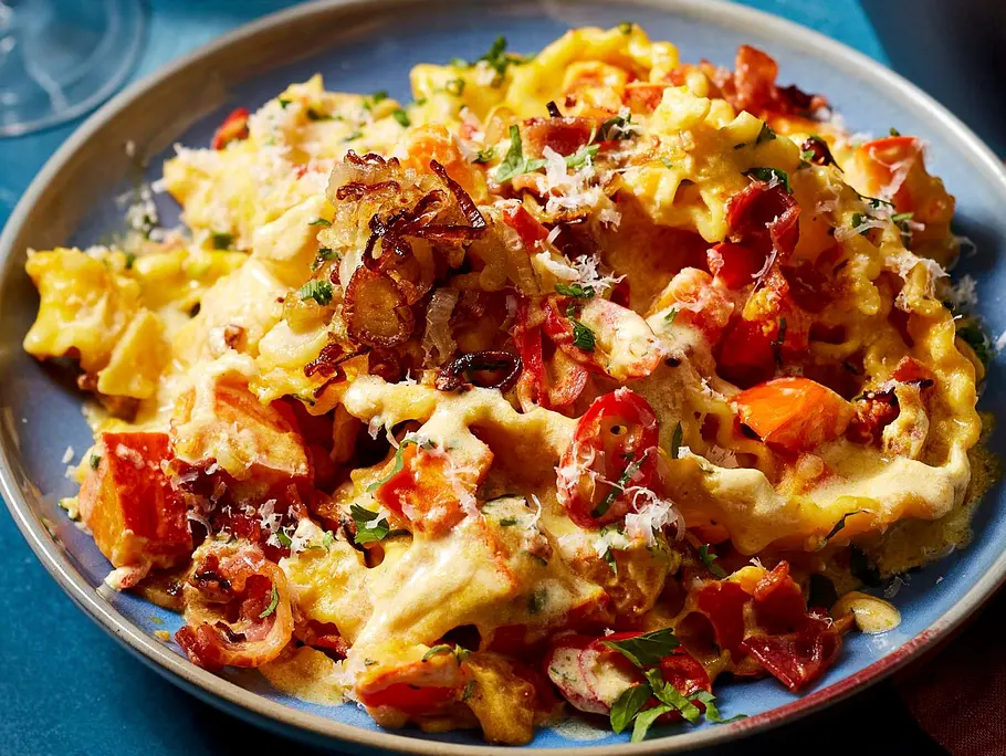 Breite Bandnudeln in cremiger Kürbissauce mit knusprigen Pancetta-Stückchen und frisch geriebenem Parmesan, serviert auf einem blauen Teller.