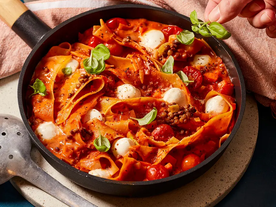 One-Pan-Lasagne mit gewellten Lasagneplatten, vegetarischer Bolognese aus Linsen und Tomaten, kleinen Mozzarella-Kugeln und Basilikum, serviert in einer schwarzen Pfanne.