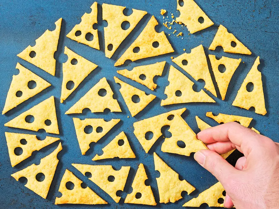Knusprige, goldgelbe Gouda-Cracker in dreieckiger Käse-Optik mit ausgestanzten Löchern, verteilt auf einer blauen, strukturierten Unterlage.