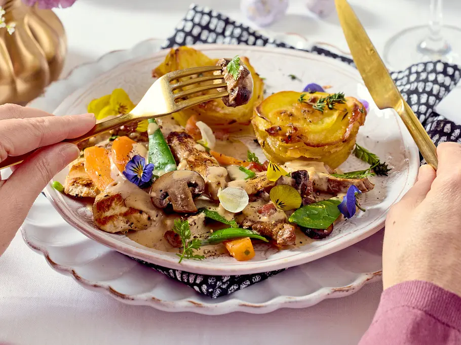 Cremiges Frühlings-Ragout mit zartem Fleisch in heller Sauce, frischem Frühlingsgemüse wie Karotten, Zuckerschoten und Champignons, garniert mit essbaren Blüten, serviert auf einem weißen Teller mit goldbesteck und knusprigen Kartoffeltürmchen als Beilage.