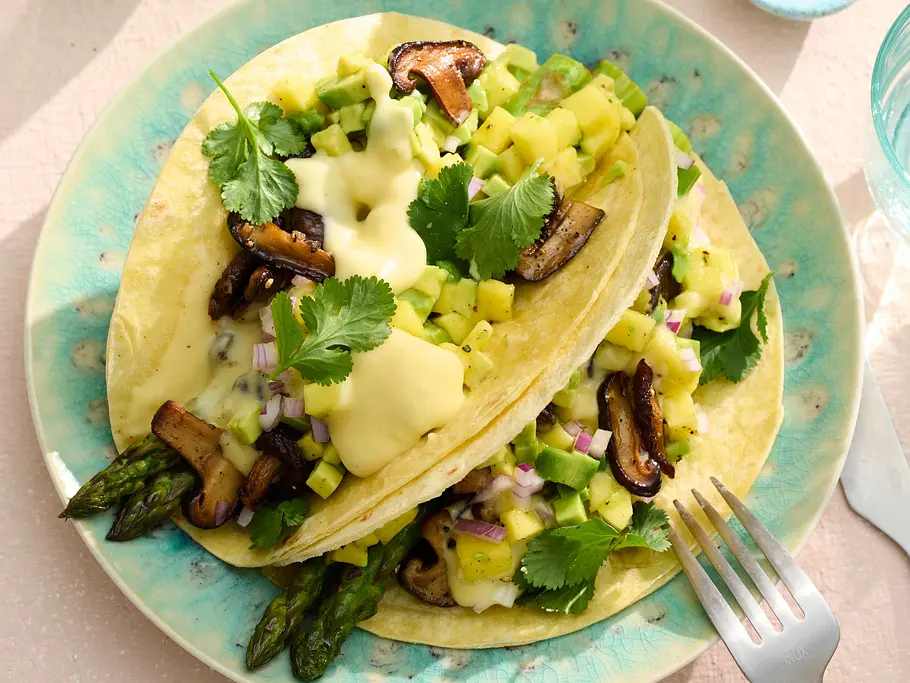 Weiche Tacos, gefüllt mit grünem Spargel und gebratenen Shiitake, getoppt mit cremiger Sauce Hollandaise, Avocadowürfeln, roten Zwiebeln und frischem Koriander, angerichtet auf türkisfarbenen Tellern.