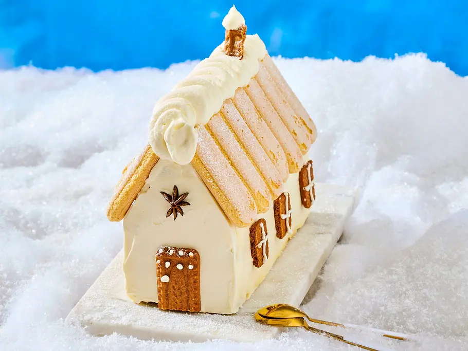 Tiramisu-Honigkuchenhaus mit Biskuitdach, verziert mit Zuckerguss und kleinen Honigkuchenfenstern, angerichtet auf einer weißen Platte mit goldener Kuchengabel.