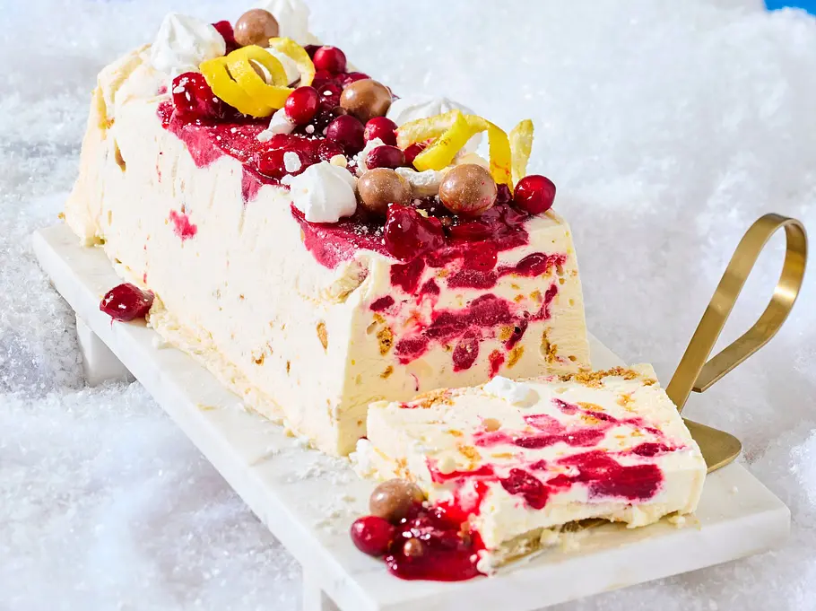 Cremiges Semifreddo in Scheiben, durchzogen und bedeckt mit leuchtend roter Cranberry-Sauce, garniert mit Zitronenzesten, kleinen Baisers und Schokokugeln, serviert auf einer rechteckigen Marmorplatte mit goldener Serviergabel.