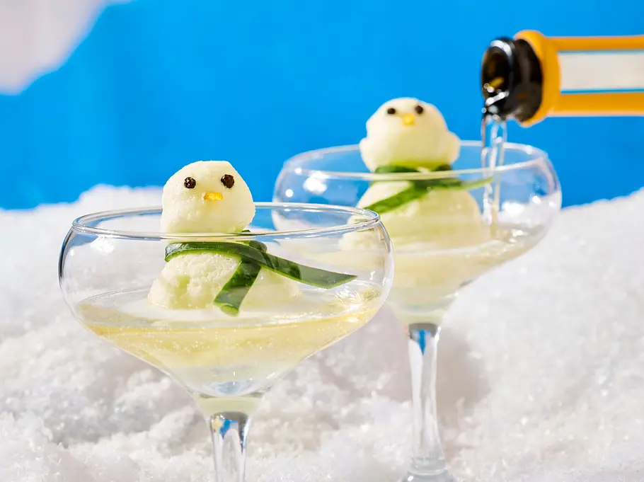 Zwei elegante Cocktailgläser mit hellem Gin-Spritz, darin je ein Schneemann aus Sorbet und Garnitur, ein Glas wird gerade mit Sprudel aufgegossen.