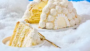Kuppelförmiger Kokos-Iglu-Maulwurfkuchen, mit Kokoskugeln dekoriert und cremiger Füllung, auf einer weißen Platte angerichtet, daneben ein angeschnittenes Kuchenstück auf einem runden Teller mit goldener Gabel. - Foto: LECKER @ Bauer Media Group